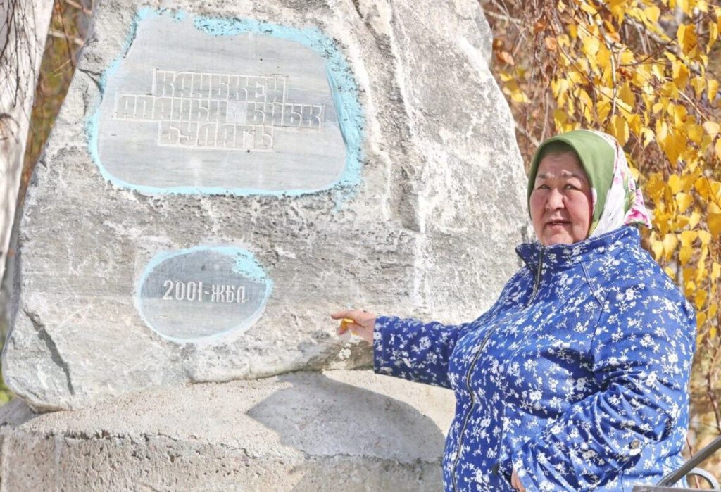 A woman wearing blue is standing by a rock with inscriptions in Kyrgyz commemorating a 2001 project